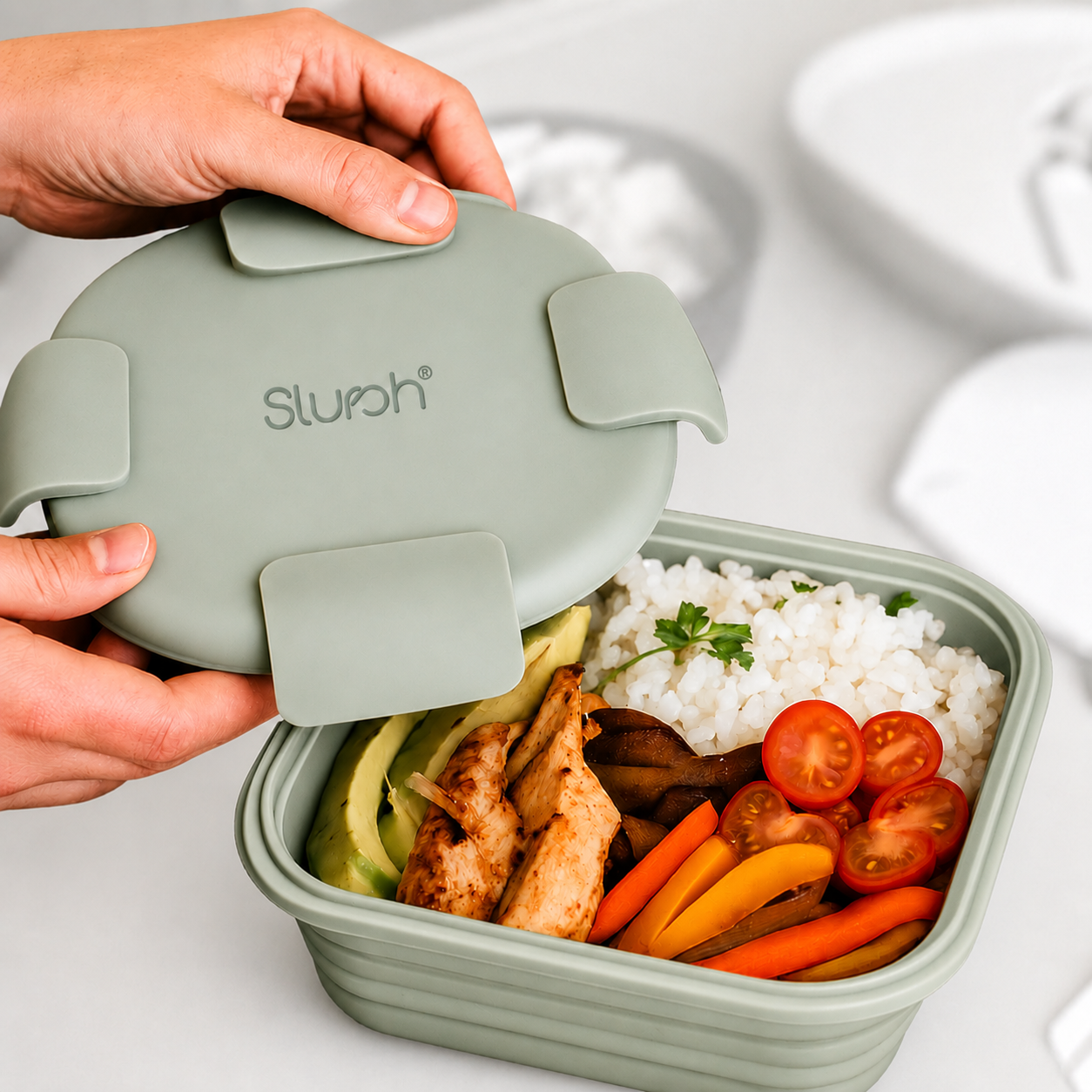 Hands opening a sage Slursh silicone box filled with rice, vegetables and sliced chicken.