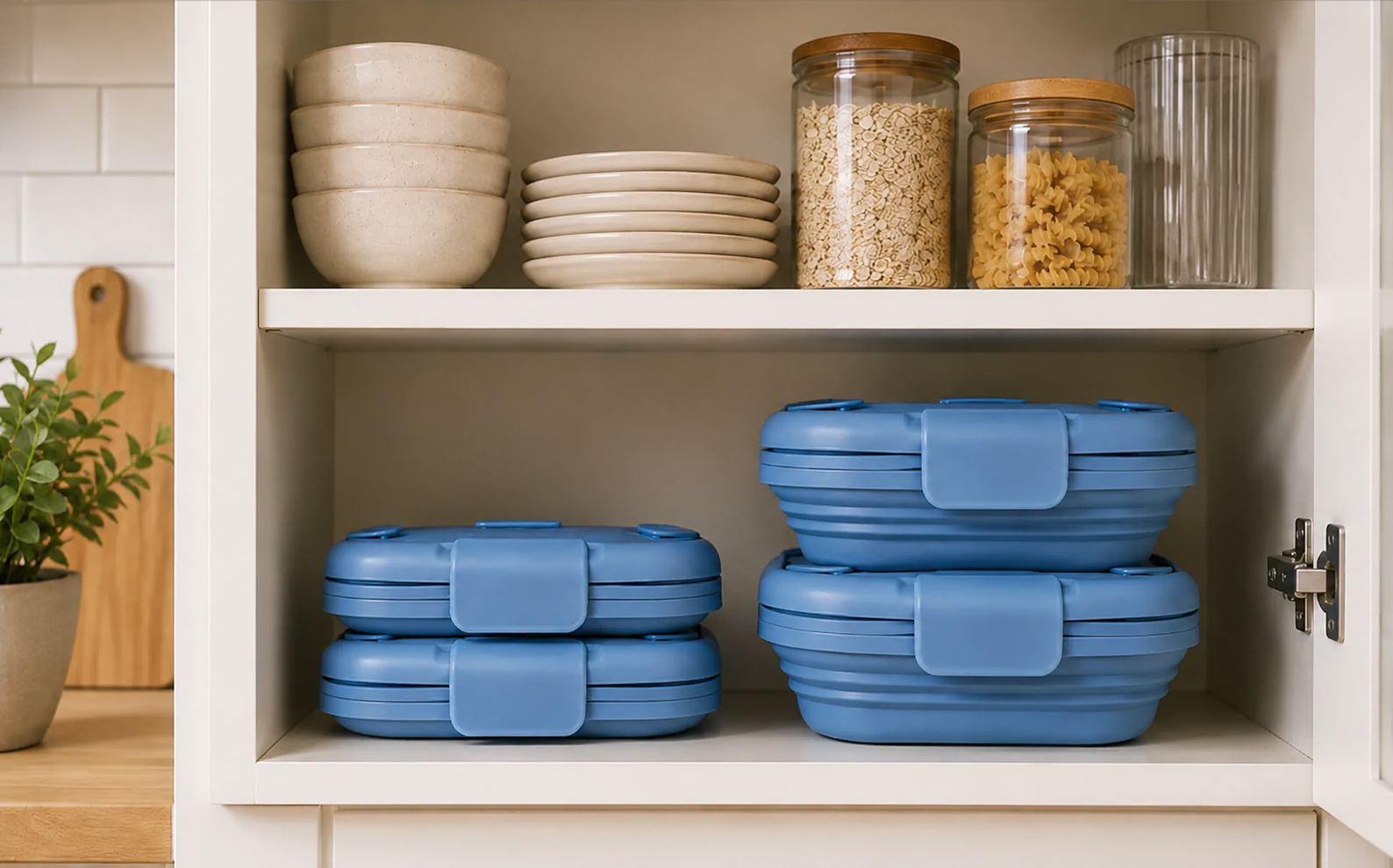 Two neat stacks of blue collapsed silicone boxes sitting inside a cream kitchen cabinet beside plates and grain jars.