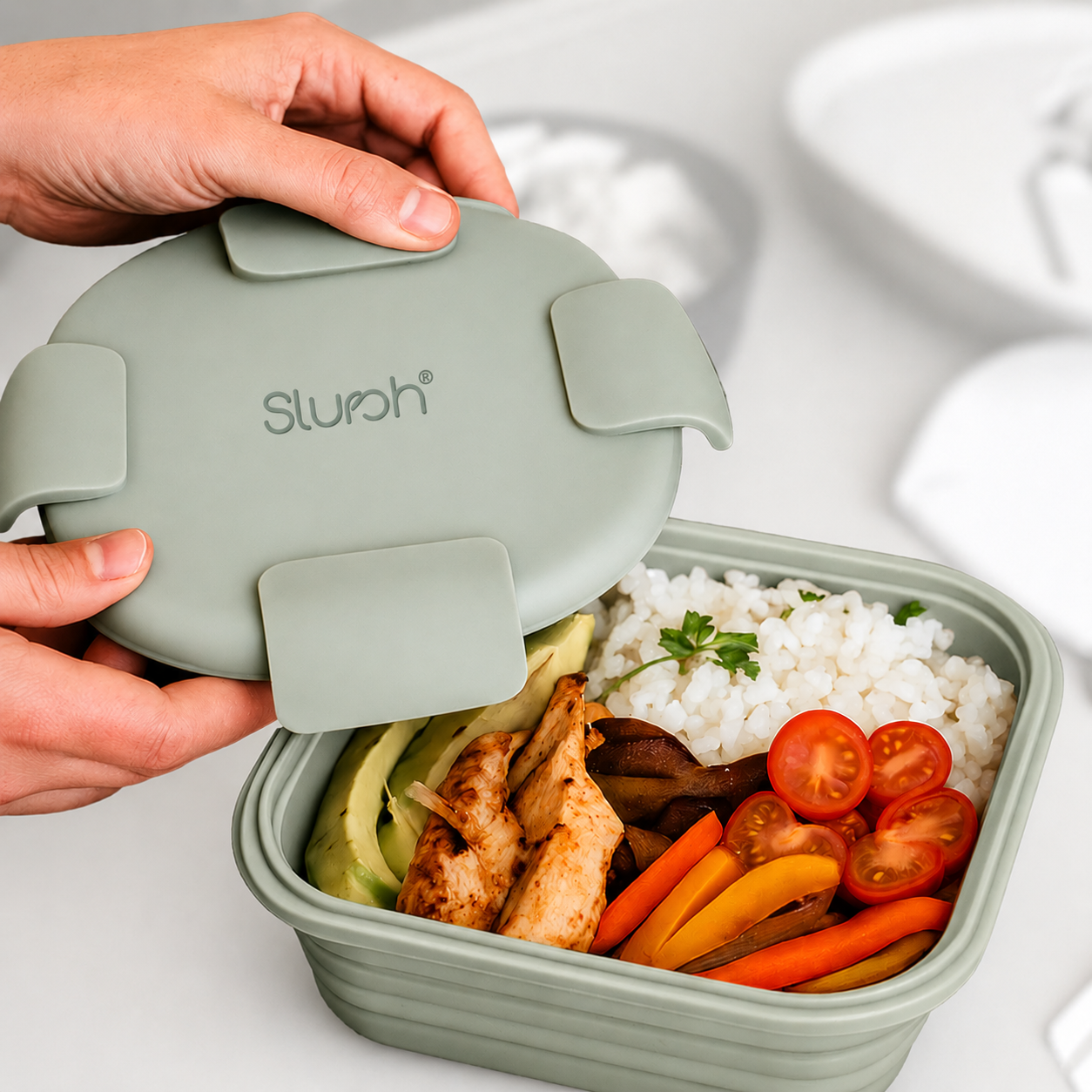 Hands lifting the sage green silicone lid off a bento style box filled with rice, grilled chicken, avocado and vegetables.